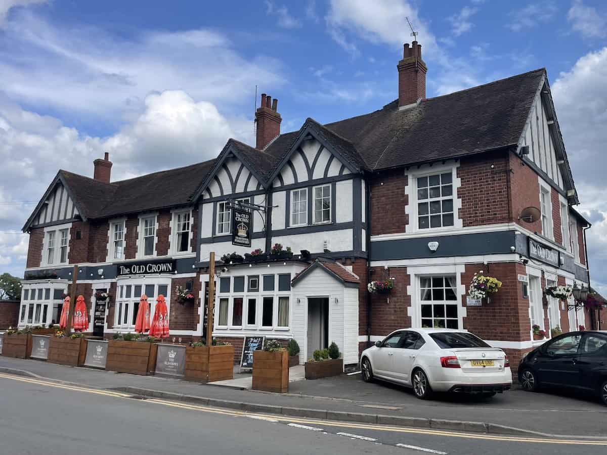 The Old Crown, a large Tudor-style pub with distinctive black and white timber framing on the upper level and red brick construction. The building has multiple chimneys, bay windows, and hanging flower baskets. Cars are parked outside and outdoor seating areas are visible.
