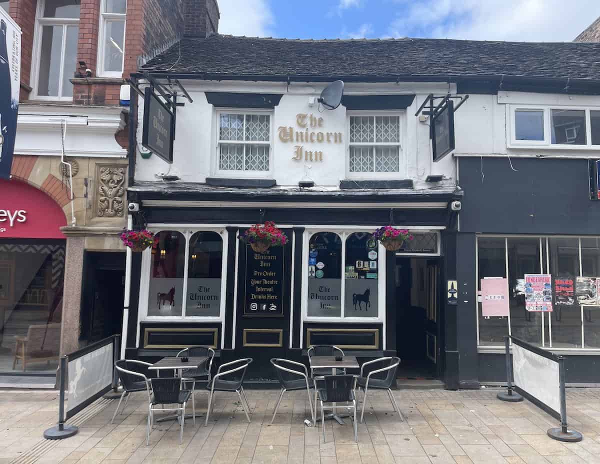 The Unicorn Inn, a traditional English pub with white-painted facade and black timber detailing, featuring outdoor seating with metal chairs and tables on the pavement. The pub has decorative windows and classic pub signage with unicorn imagery.