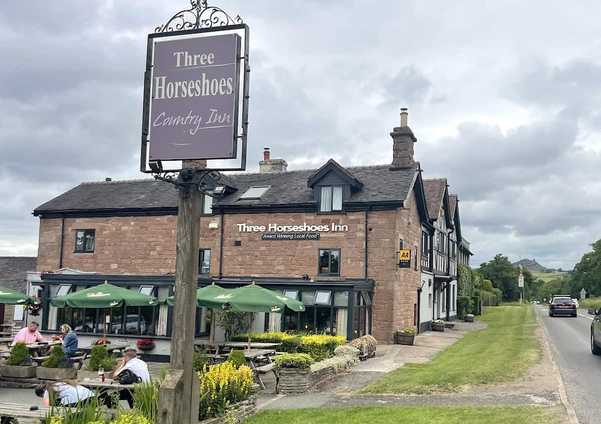 The Three Horseshoes Country Inn, showing a stone-built pub with Tudor-style timber framing. The building has a large pub sign, outdoor seating area with green umbrellas, and patrons dining at picnic tables. The setting appears rural with open countryside visible in the background.