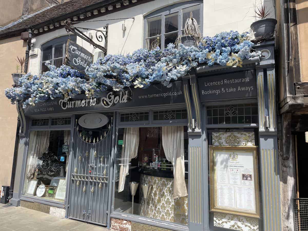 Turmeric Gold, an Indian restaurant housed in a historic timber-framed building with ornate decorative details. The storefront features abundant blue hydrangea flowers cascading from window boxes, creating a striking floral display against the traditional Tudor-style architecture.