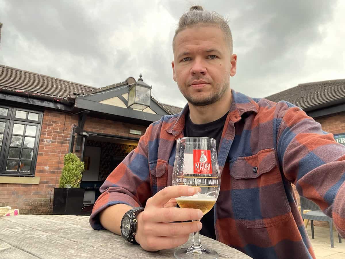 A man in a plaid shirt holds a glass of beer with a "Madri" logo while sitting at an outdoor table in front of a brick house with a modern glass extension.