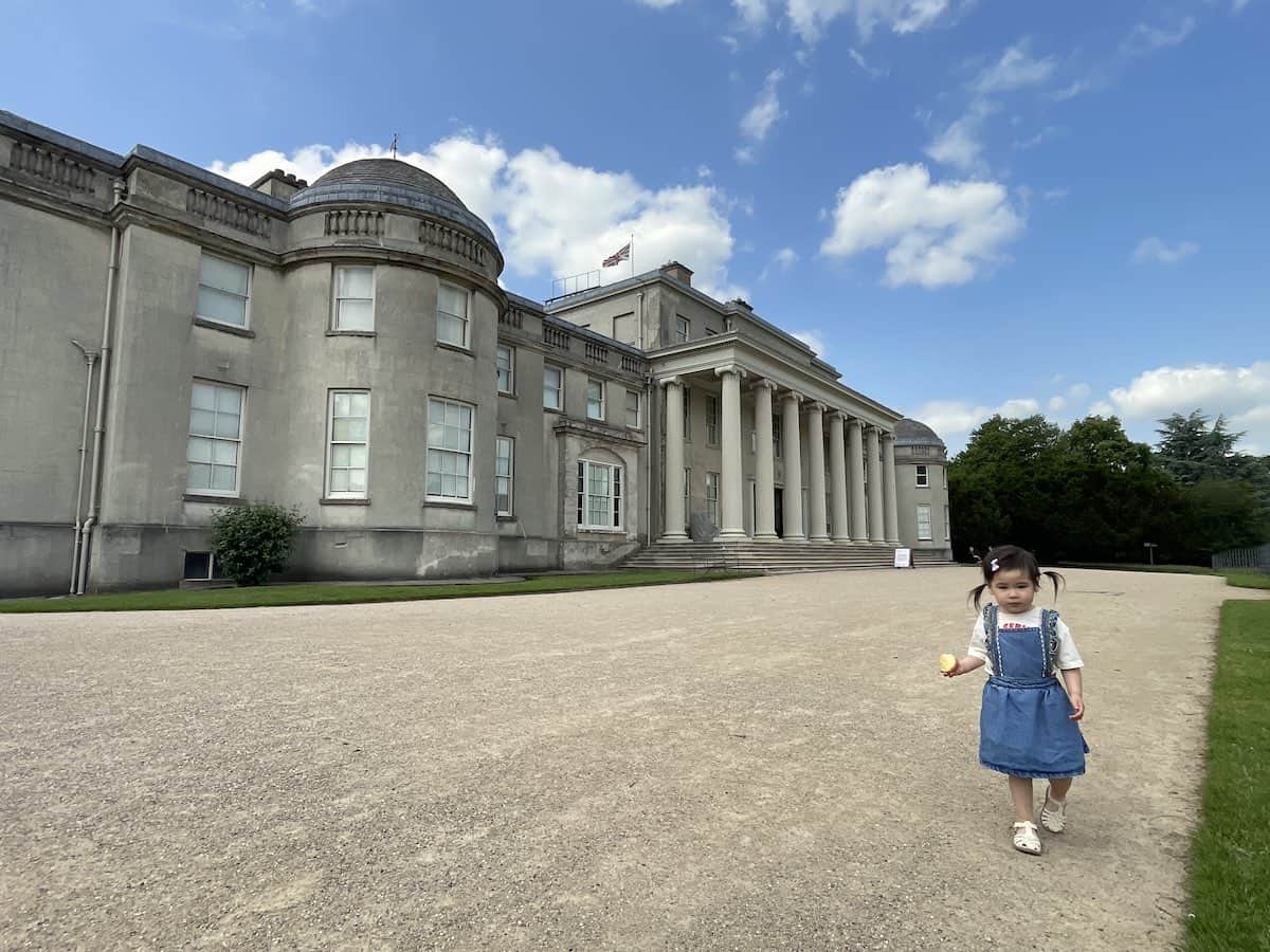 a large stately home with roman style pillars at the entrance, while a young toddler with pigtails walks towards the camera while eating an apple, at the forefront on the right side.