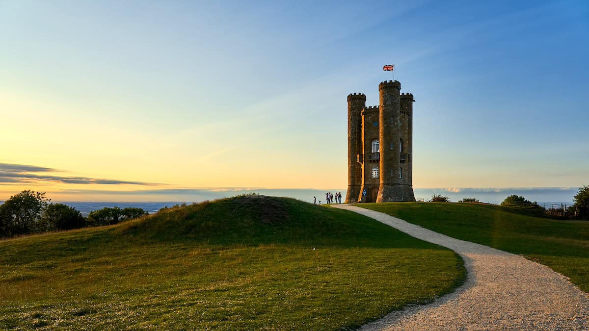 Broadway Tower photographed during golden hour, silhouetted against a dramatic sunset sky with the Union Jack flag flying from its highest point, approached by a winding gravel path across the grassy hilltop.