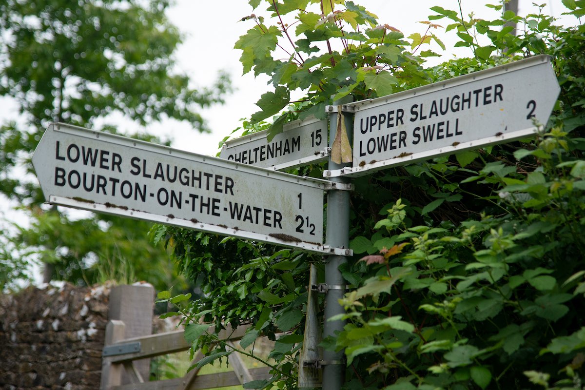 Road sign pointing to Upper Slaughter (2 miles), Lower Swell (2 miles), Lower Slaughter (1 mile) and Bourton-on-the-Water (2.5 miles) on a quiet Cotswolds country lane, surrounded by green hedgerows.