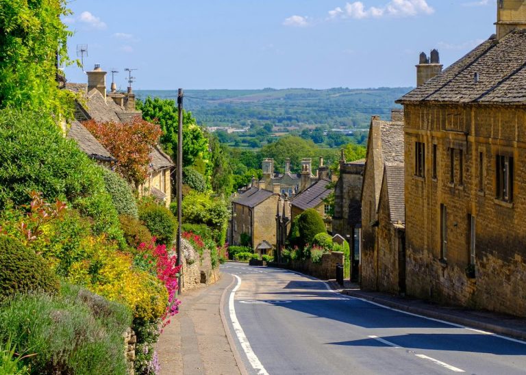 a narrow road windign down hill with honey-coloured, traditional country cottage-style buildings lining one side (the right) and shrubbery lining the other side (left), with a vast countryside spanning the horizon in the distance.