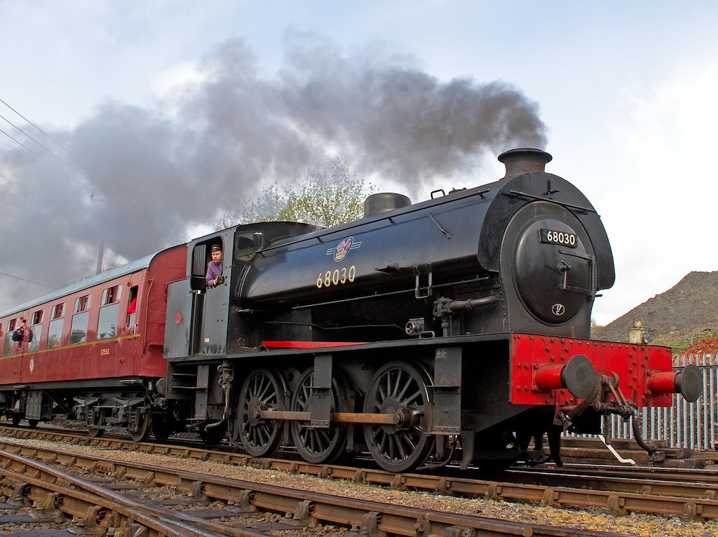 close up of a black and red steam locomotive on a railway track with black smoke billowing from its chute and people onboard looking out the windows