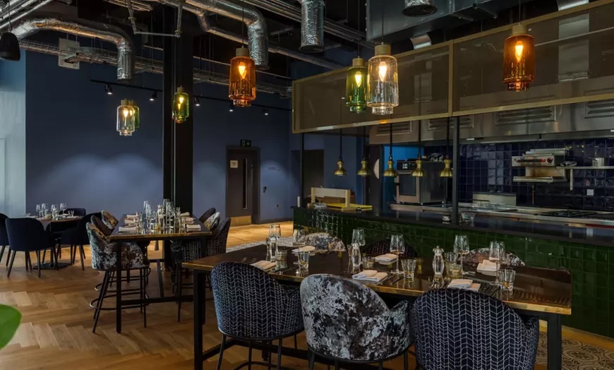 dark large restaurant room with a bar and dining tables with blue upholstered chairs and glasses on the tables, with fancy glass lighting