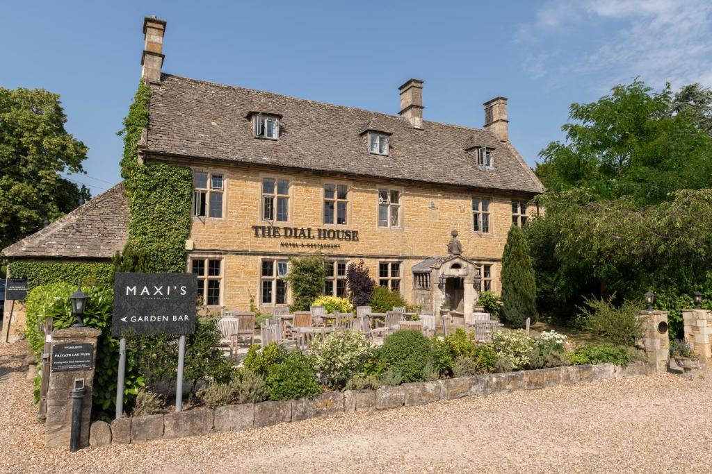 A honey-coloured countryside hotel called the Dial House with an outdoor dining area, and surrounded by some trees, wall-creeping florals and small hedges and florals at the front, with a rustic sign for The Dial House, featuring "MAXI'S" and "GARDEN BAR."
