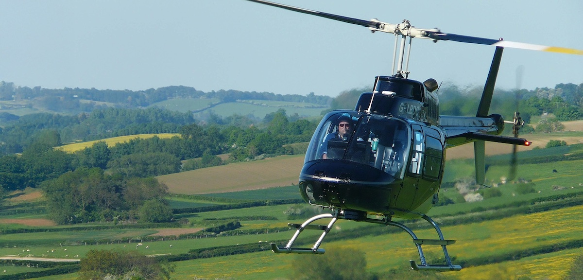 view of a small black helicopter flying over an English countryside, with a pilot visible in the cockpit.