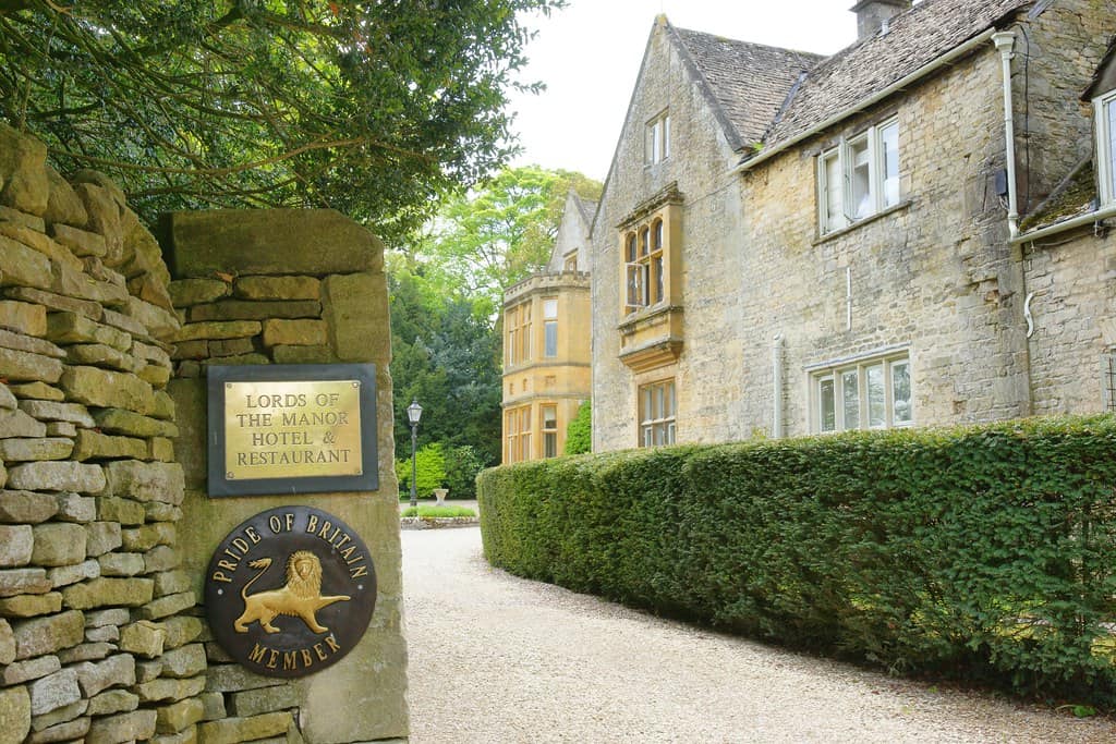 An old country manor house surrounded by hedges, with a sign for "Lords of the Manor Hotel & Restaurant." The text is elegantly formatted, with "LORDS OF" above "THE MANOR," and "HOTEL & RESTAURANT" below.