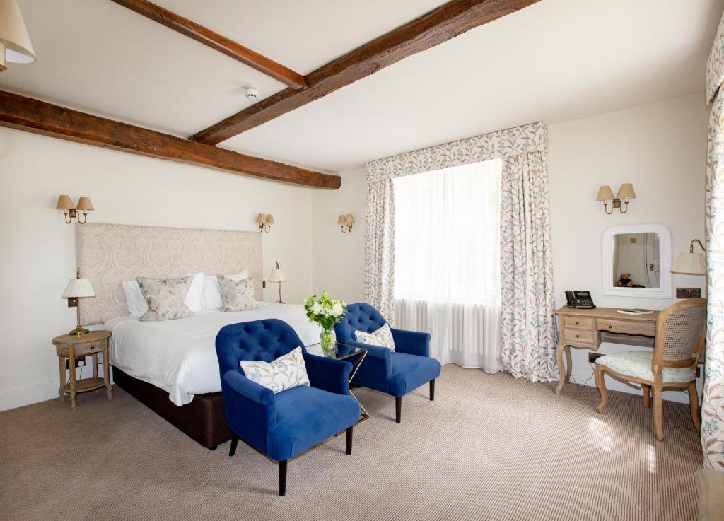 large cream bedroom with timber beams on the ceiling, white double bed with cushions and a cream headboard, blue suede armchairs infrotn of the bed, and a wooden dressing table with wall mounted mirror, in the Lords of the Manor Hotel in Upper Slaughter, a stunning honey-stone Cotswolds country house with elegant windows and a classic Georgian facade, set within beautiful green grounds.