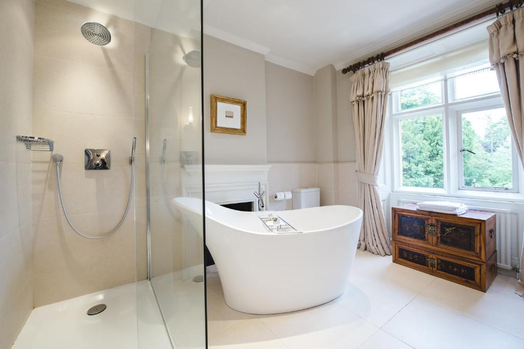 large cream bathroom with modern white isolated bathtub, walk in shower with glass screen, and bright windows with long curtains tied back, in the Lords of the Manor Hotel in Upper Slaughter, a stunning honey-stone Cotswolds country house with elegant windows and a classic Georgian facade, set within beautiful green grounds.