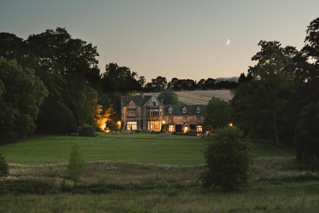 Lords of the Manor Hotel after sunset in Upper Slaughter, a stunning honey-stone Cotswolds country house with elegant windows and a classic Georgian facade, set within beautiful green grounds. You can see the lights on inside the building and a crescent moon visible on the darkening sky.