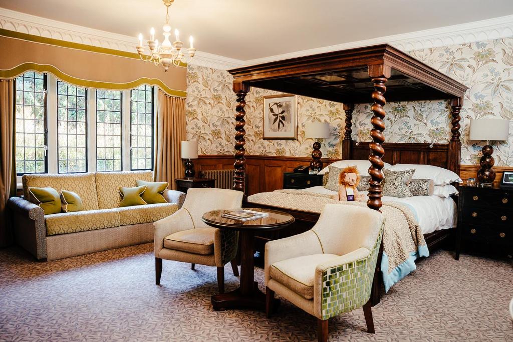 A luxurious Edwardian style bedroom with wooden four-post bed, floral wallpaper with cream and beige colours throughout the room, two cream chairs at the foot of the bed with a round table, and a sofa by the window.