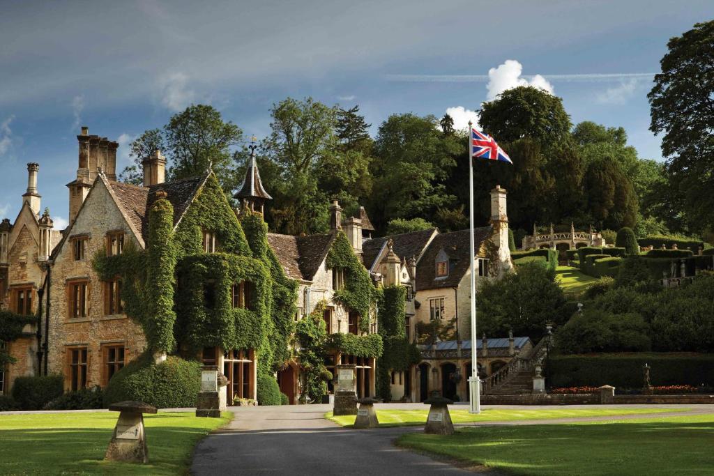 A view of the stone-built Manor House hotel in the Cotswolds, surrounded by foliage with neat lanws in front and large trees behind, and a pole in front with a union jack flag waving from it.