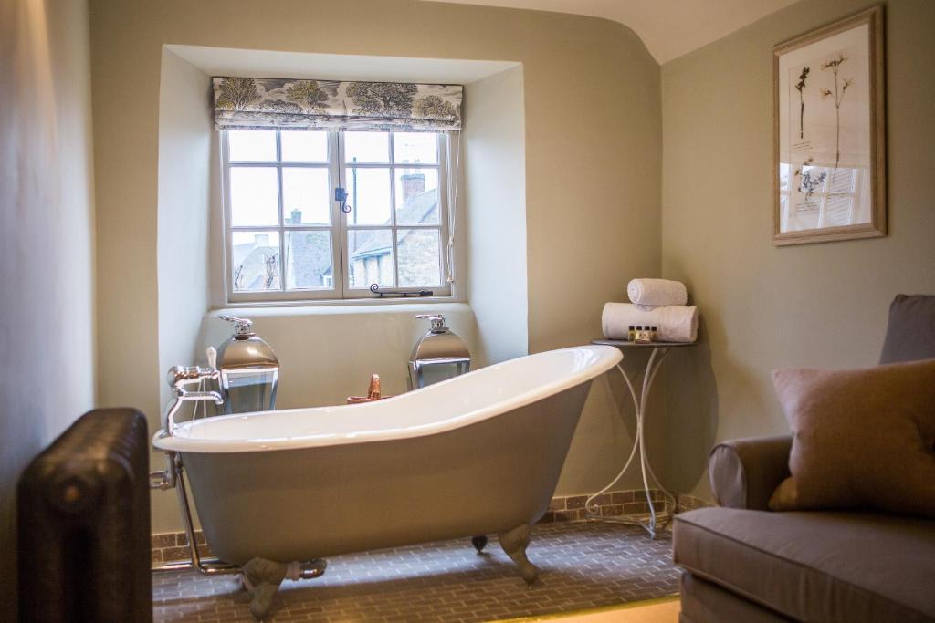 cream bathroom with a modern isolated white bathtub, brown sofa, small window with floral blinds rolled up, and white towels on a side table, at the Porch House in Stow-on-the-Wold, England's oldest inn.