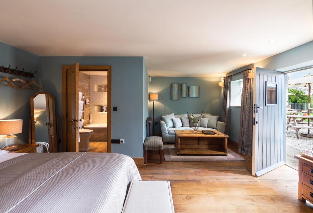 A modern design hotel bedroom with king size bed with en-suite bathroom in the corner and a door open leading to the garden. Oak flooring, blue decor, and a sofa sits in another corner with a wooden table.