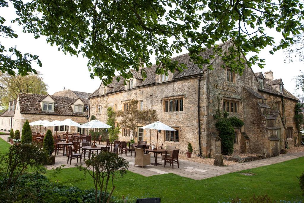 A view of the stone-built Slaughter's Inn in the Cotswolds, seen from the rear under the shade of a large tree whose branches arch over the top of the scene.