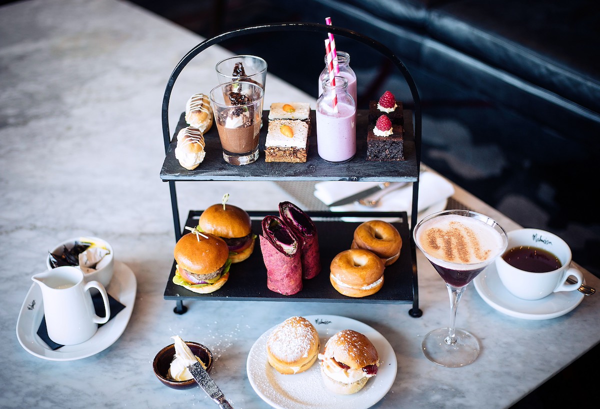 Modern two-tier black slate afternoon tea stand featuring mini burgers, beetroot wraps, glazed donuts, eclairs, chocolate mousse, layered cake bar, and pink milk bottle, served with scones, coffee, and cocktails