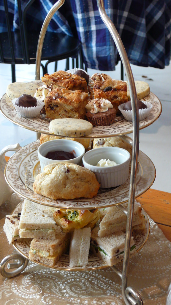 Traditional three-tier afternoon tea stand with ornate floral china plates, featuring finger sandwiches on bottom, scone with jam and cream on middle tier, and assorted cakes including shortbread cookies, chocolate truffles, and frosted cupcakes on top
