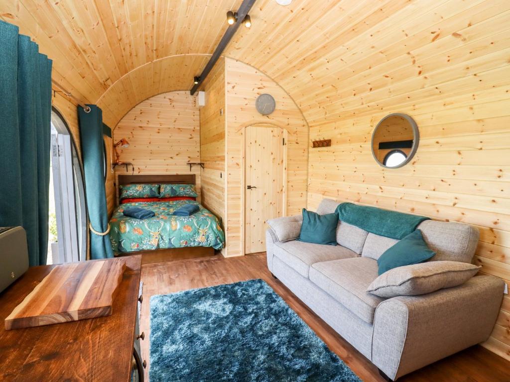 Cozy interior of wooden glamping pod with curved pine walls and ceiling, featuring double bed with teal floral bedding in alcove, gray sofa with teal cushions, blue shag rug, round porthole window, fold-down wooden table, and teal curtains