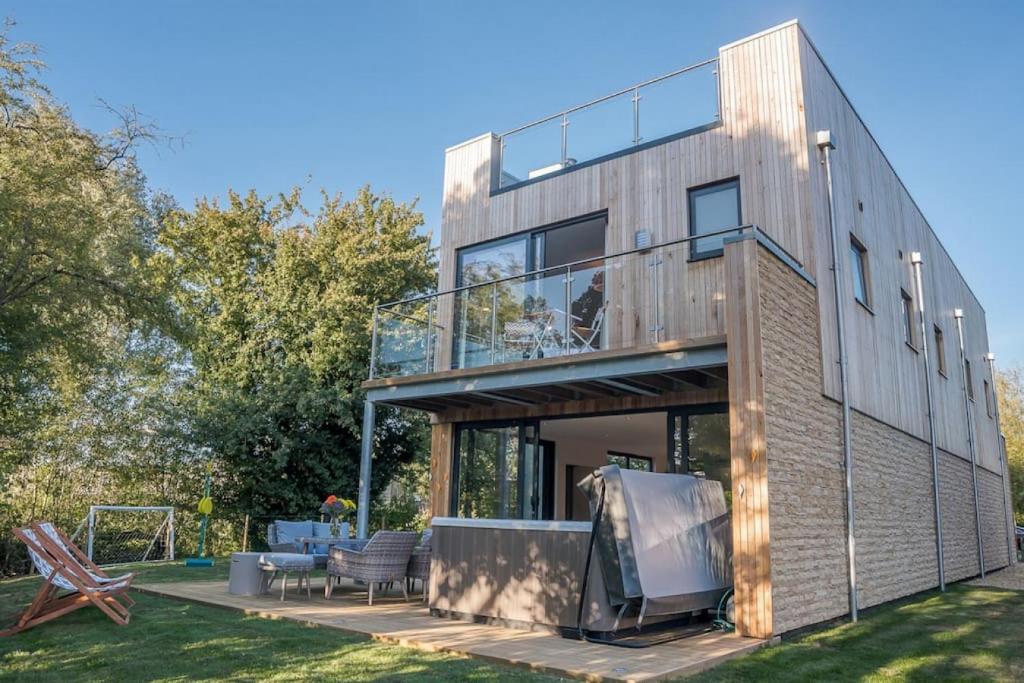 Modern two-story vacation home with vertical wood and beige horizontal siding, covered hot tub on ground level patio with wicker outdoor furniture and deck chairs on lawn, glass-railed upper balcony with white furniture, floor-to-ceiling windows, clear blue sky
