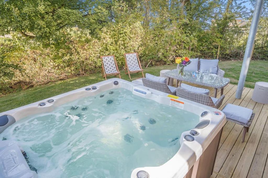 Large white hot tub with jets on wooden deck beside gray wicker outdoor dining set with white cushions, two wooden deck chairs on lawn, flower arrangement on table, garden with hedges and trees in background