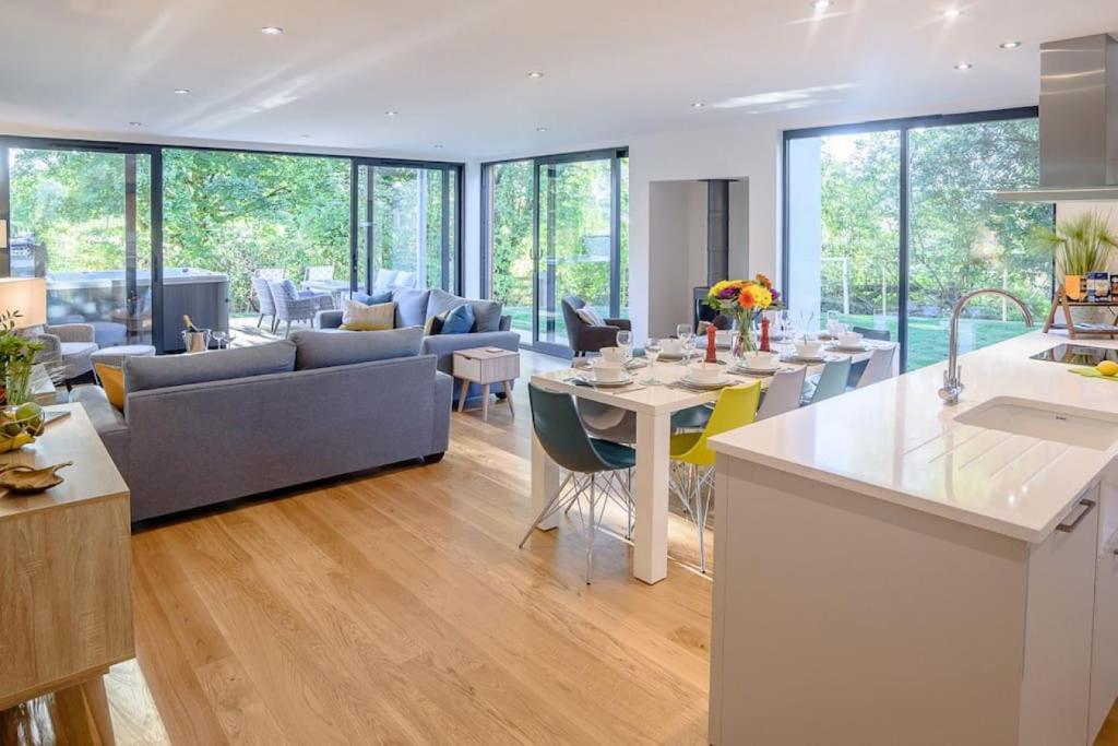 Spacious open-plan living area with light wood flooring, gray L-shaped sofas, white dining table with colorful Eames-style chairs, white kitchen island with breakfast bar, floor-to-ceiling glass doors and windows overlooking garden with patio furniture, modern pendant lighting