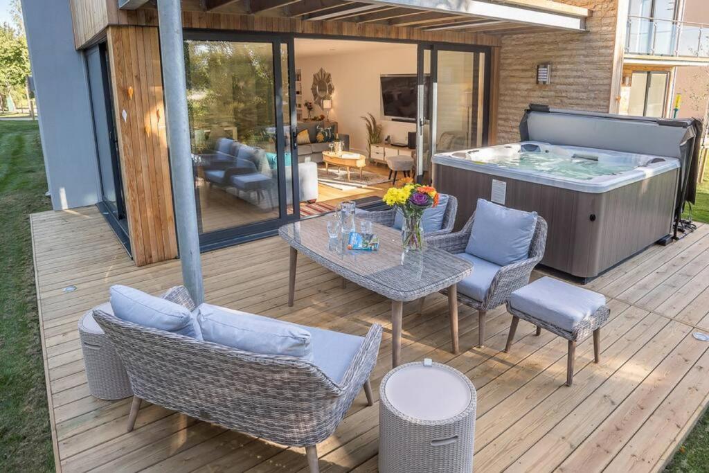 Wooden deck with gray wicker outdoor sofa and dining set with blue cushions, hot tub with open cover, floor-to-ceiling sliding glass doors opening to modern living room with gray furniture and wall-mounted TV, vertical wood and stone exterior cladding, covered pergola overhead