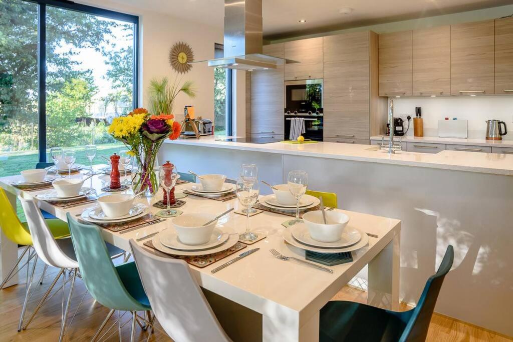 Modern kitchen and dining area with white glossy breakfast bar set for eight with white dishes and colorful chairs in yellow, teal, white and green, light wood handleless cabinets, stainless steel appliances and range hood, sunburst mirror, large windows with garden views