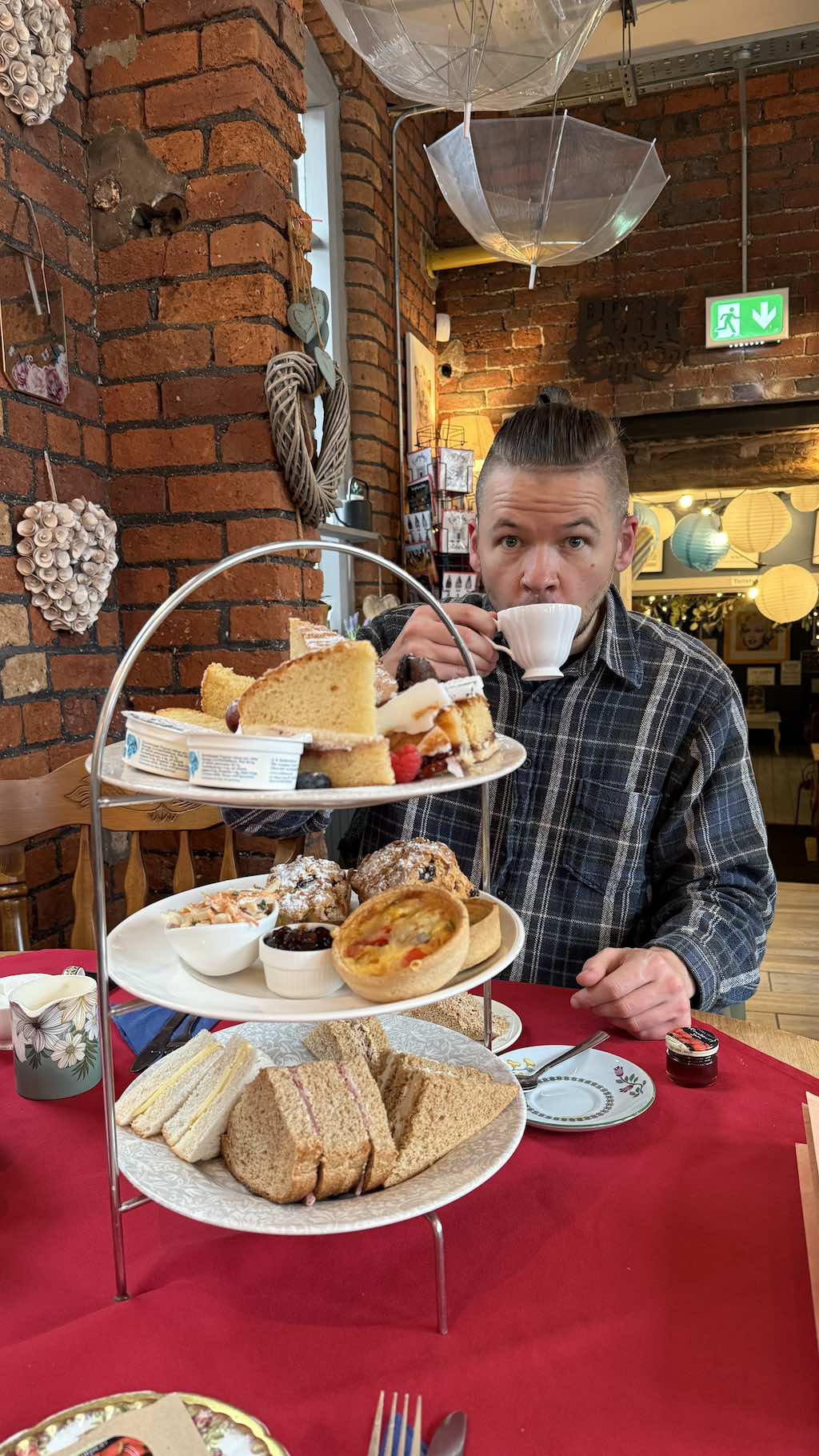 Person in plaid shirt sitting at table with elaborate three-tiered afternoon tea stand featuring sandwiches, scones, cakes, and pastries, holding teacup in rustic brick-walled café with decorative lighting