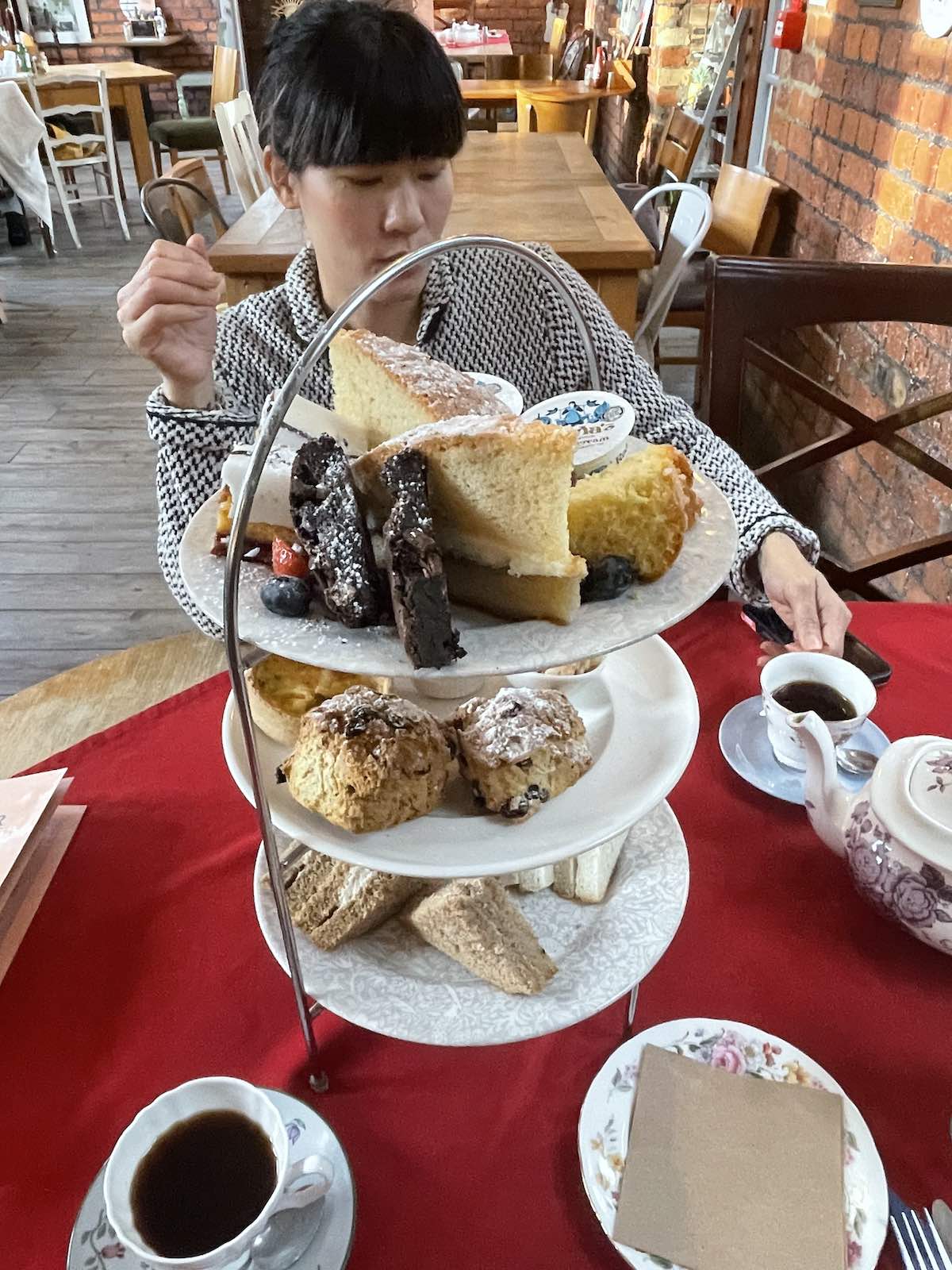 Person in patterned sweater reaching for food on three-tiered afternoon tea stand filled with cakes, scones, brownies, and finger sandwiches, with teacups on table in café with exposed brick walls