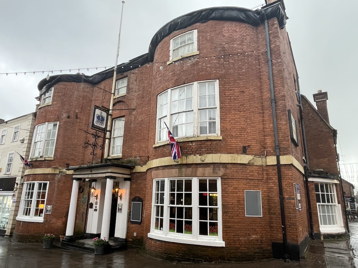 Historic red brick building with distinctive curved turret architecture featuring large white-framed windows, white columned entrance portico, Union Jack flag hanging from facade, pub sign mounted on wall, and string lights along roofline on overcast day with wet pavement