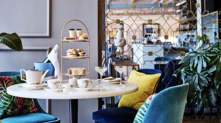 Luxurious afternoon tea setup on white marble table with gold three-tier stand displaying scones, finger sandwiches, and colorful petit fours, accompanied by champagne coupes and ornate teacups, in stylish lounge with velvet chairs in blue, yellow and teal, tropical print cushions, and geometric gold mirror wall