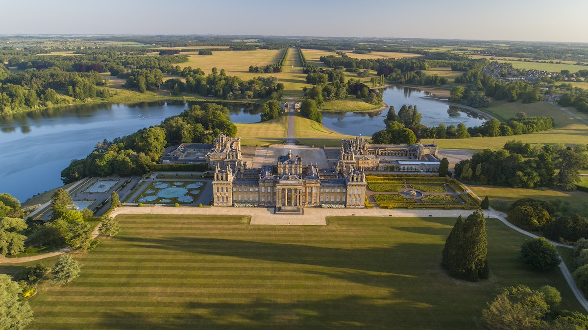 Aerial view of Blenheim Palace, grand Baroque country house surrounded by landscaped parkland with ornamental lakes, formal gardens with geometric patterns, long tree-lined avenue extending into distant countryside, expansive green lawns with striped mowing patterns, reflecting pools, wooded areas