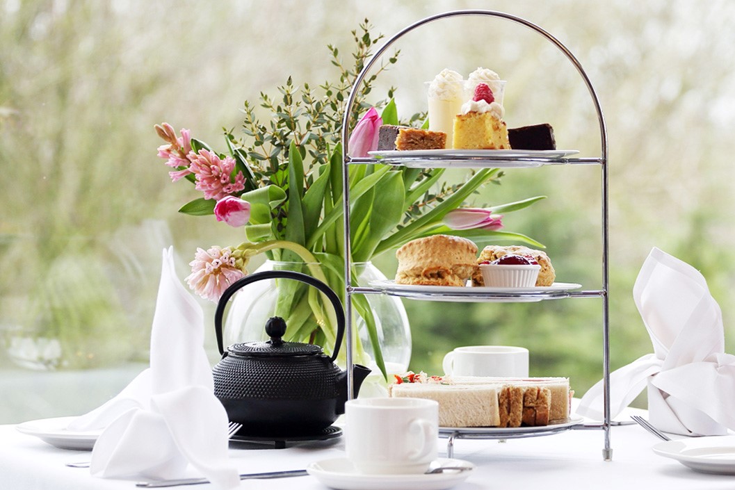 Elegant chrome three-tier afternoon tea stand with finger sandwiches on bottom tier, scones with jam and clotted cream on middle tier, colorful petit fours and cakes on top tier, white china teacup and saucer, black cast iron teapot, tall glass vase with pink tulips and spring flowers, white folded napkins, bright window background