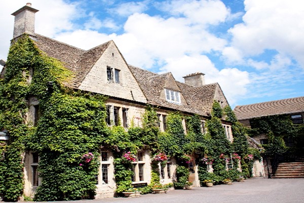 Historic Cotswold stone manor house hotel with steep pitched roof and multiple gables, walls covered in climbing green ivy with pink hanging flower baskets, mullioned windows, stone chimney, courtyard entrance, blue sky with white clouds