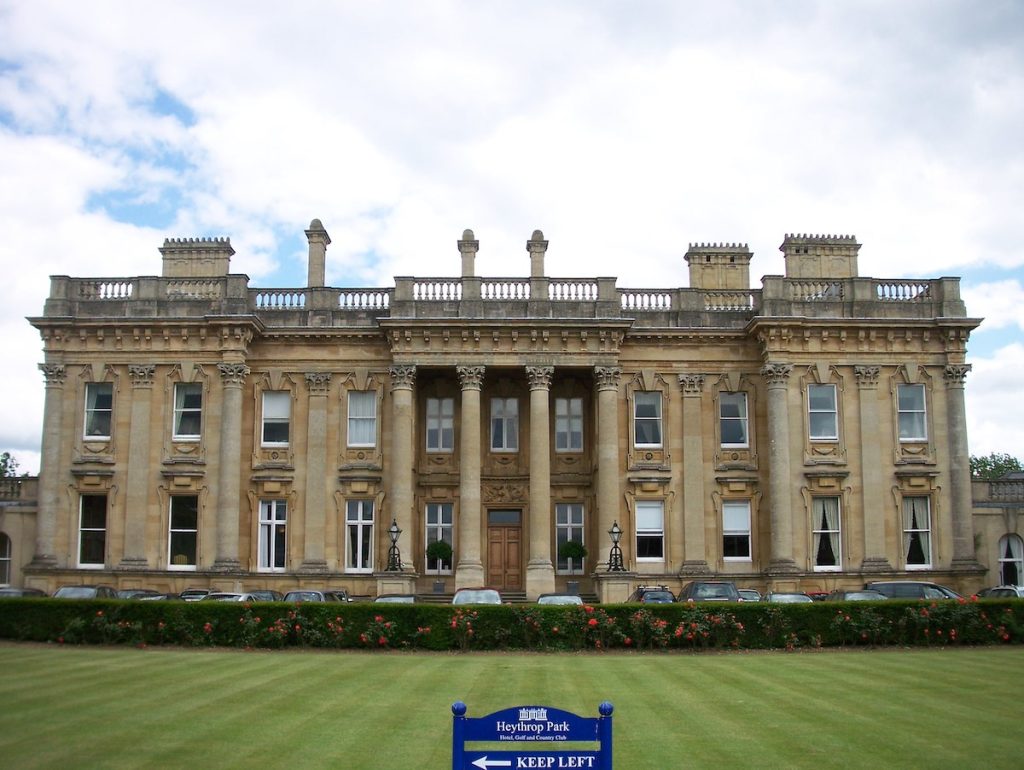 Grand neoclassical Heythrop Park mansion with honey-colored stone facade, tall Corinthian columns flanking central entrance, ornate balustraded roofline with decorative chimneys, multiple symmetrical windows, manicured lawn with flower beds in foreground, blue directional sign reading 'KEEP LEFT'