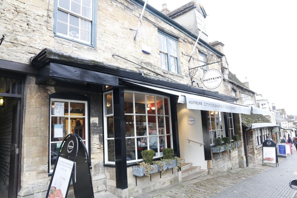 Historic Cotswold stone building with Huffkins bakery and tearoom storefront featuring black-painted woodwork, multi-pane windows, A-frame sidewalk sign, window boxes with plants, cobblestone street, row of shops along traditional English village high street