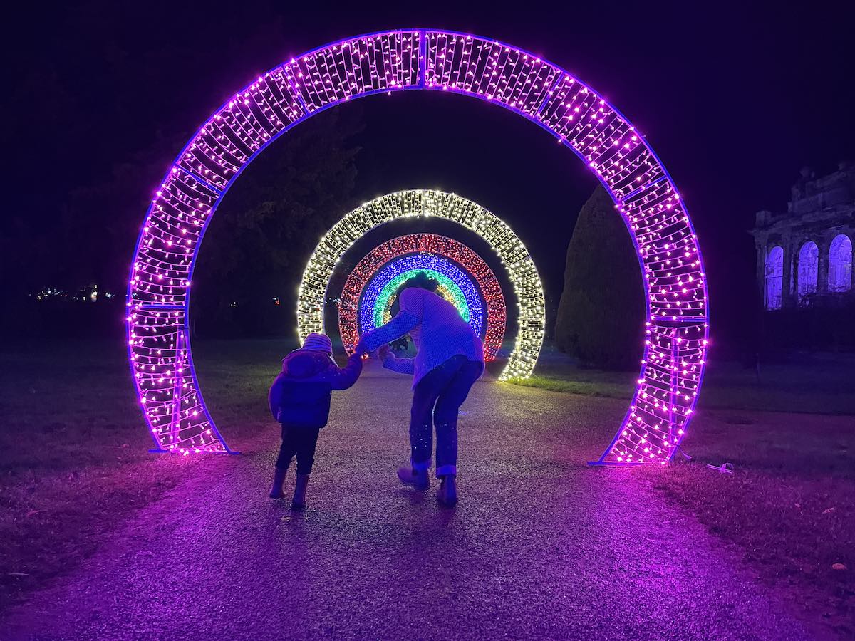 Adult and child holding hands walking through illuminated archway tunnel made of concentric rings of LED lights in purple, white, copper, blue, and green at nighttime winter light festival, with historic abbey ruins visible in background