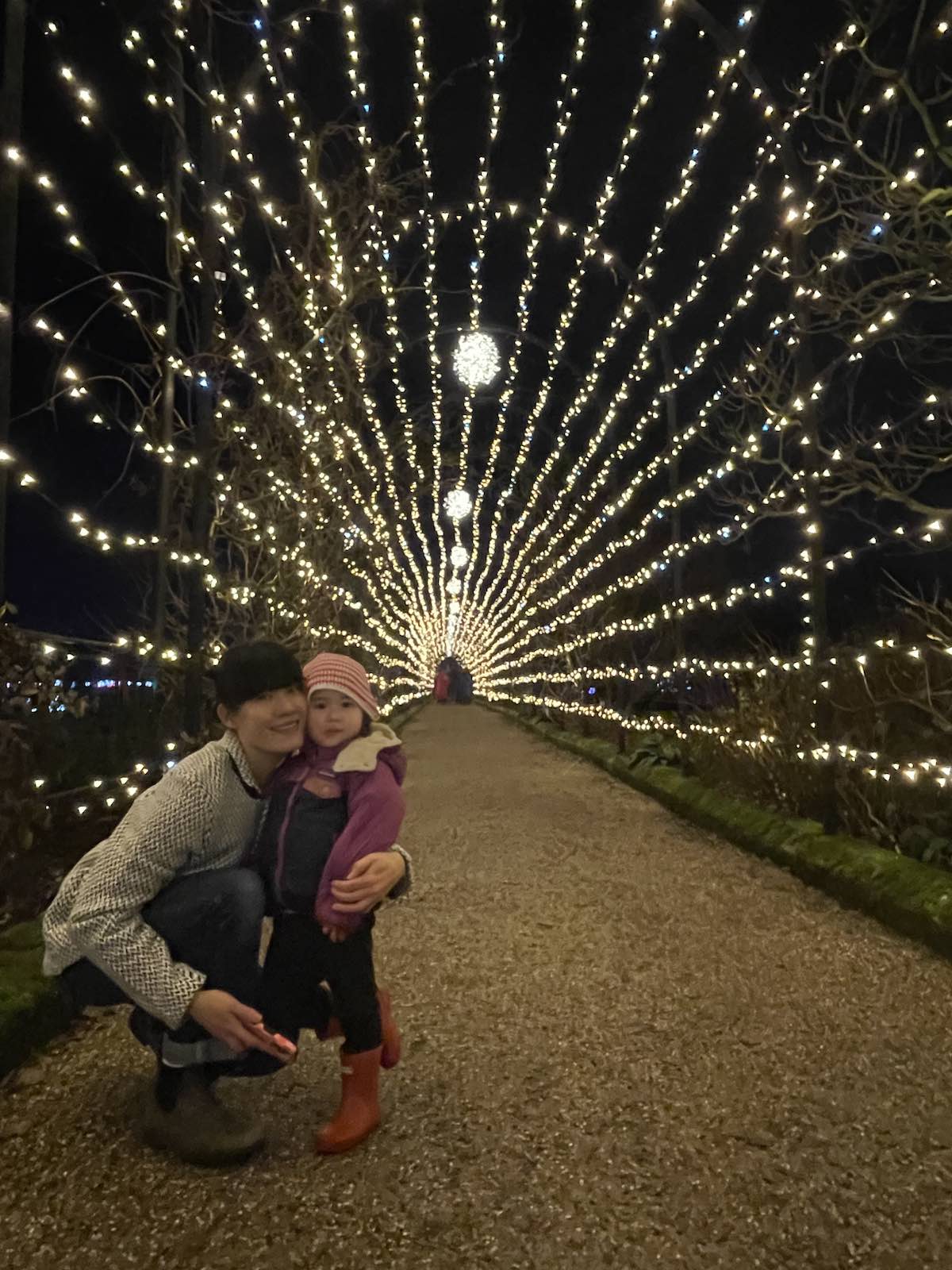 Mother and young daughter in winter clothing kneeling on gravel path beneath tunnel of warm white fairy lights radiating outward in peacock or sunburst pattern through bare tree branches at nighttime Christmas light display