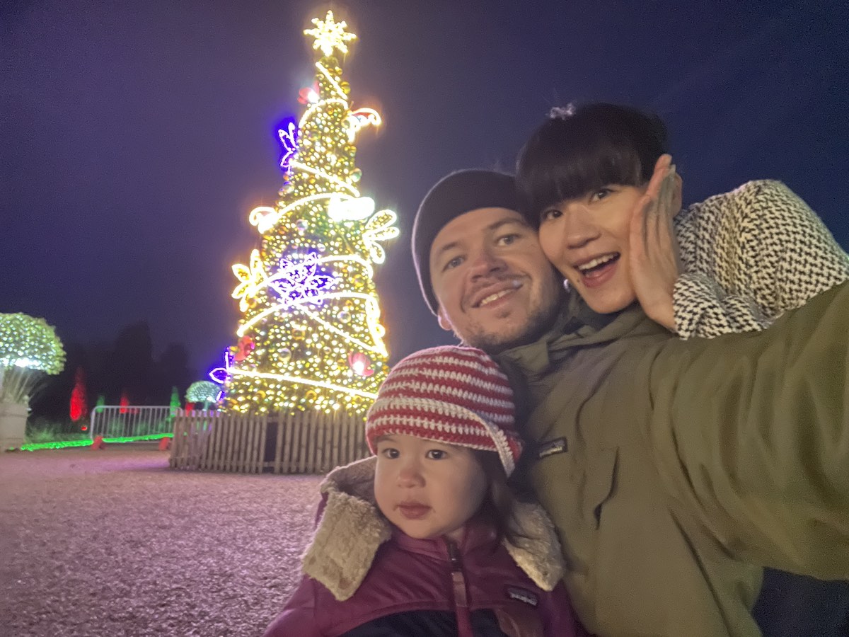 Nighttime family selfie of two adults and young child in winter coats posing in front of illuminated Christmas tree decorated with warm white fairy lights and colorful light-up animal shapes including butterflies and birds, with green spherical light sculptures in background
