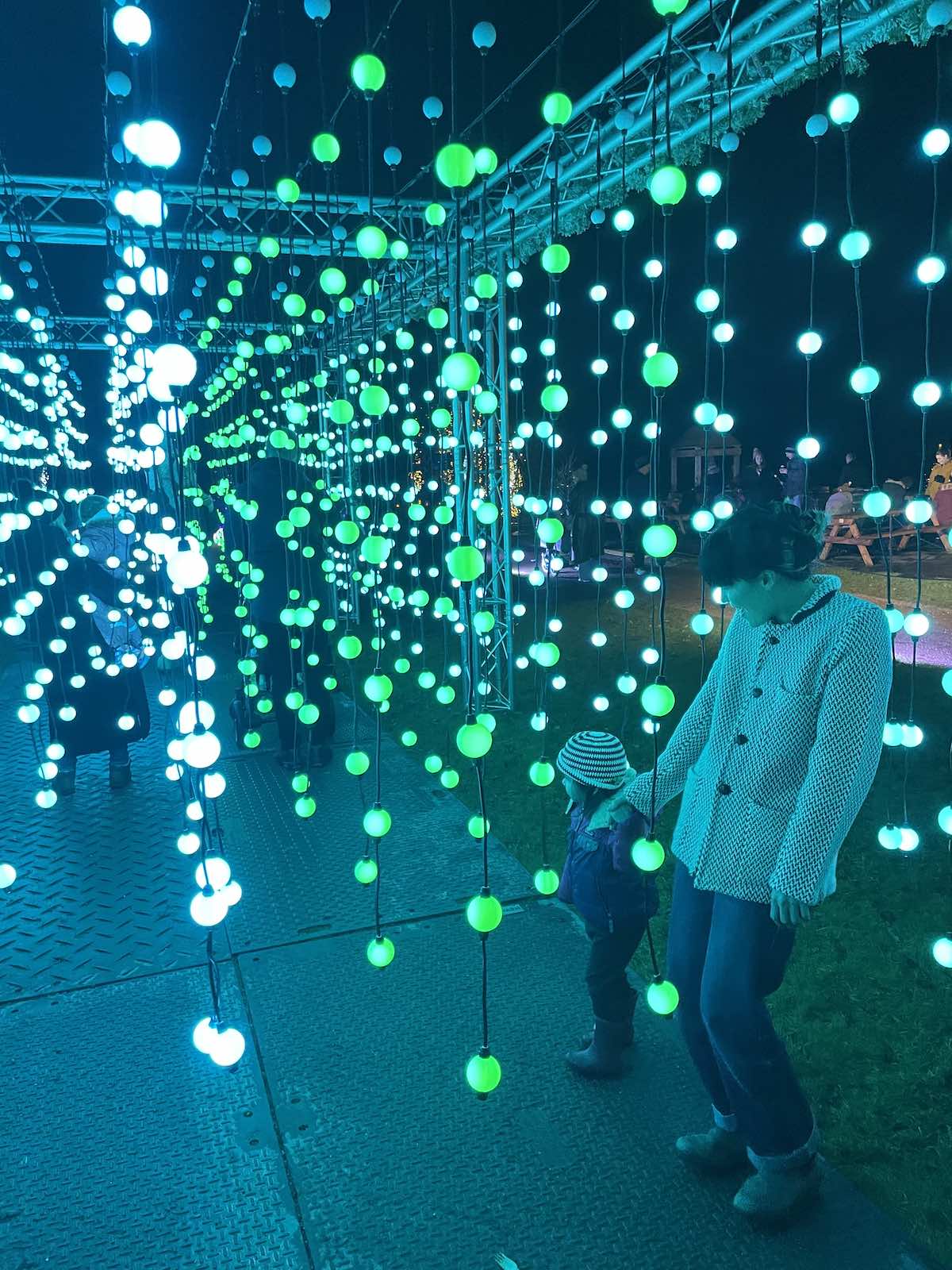 Person in textured coat and striped knit hat standing among immersive light installation with hundreds of suspended glowing orbs in white, cyan, and green forming curtains of light under metal framework structure at nighttime outdoor event
