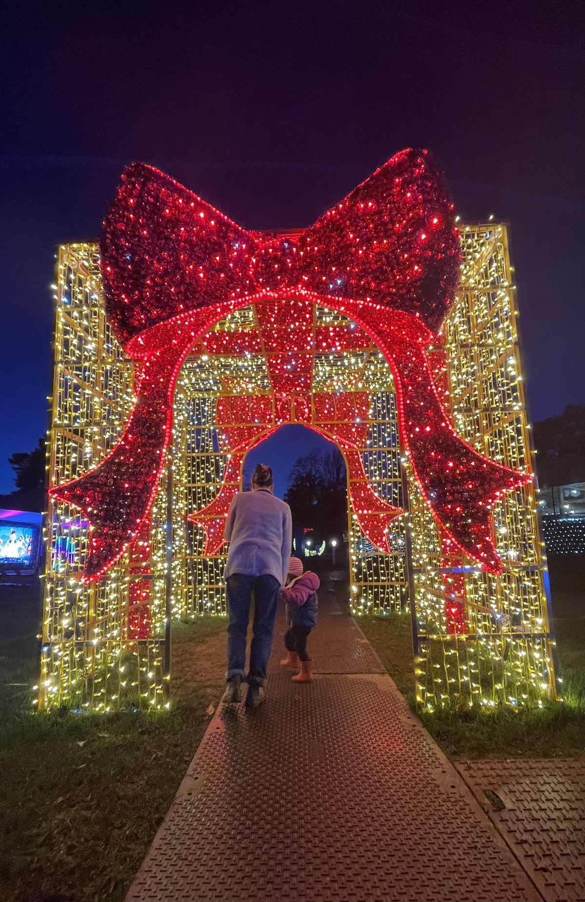 Parent and child walking on lit pathway through illuminated Christmas present archway structure decorated with red lights forming giant bow on top and golden yellow curtain lights on sides against dark evening sky at outdoor winter festival