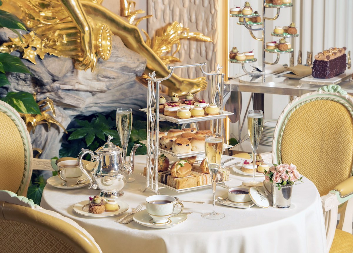 Luxurious afternoon tea table set with white linens and yellow upholstered chairs, featuring chrome three-tier stand with scones, finger sandwiches and pastries, silver teapot, champagne flutes, fine china teacups, pink roses, ornate gold swan wall sculpture, and cupcake display tower in elegant cream and gold dining room