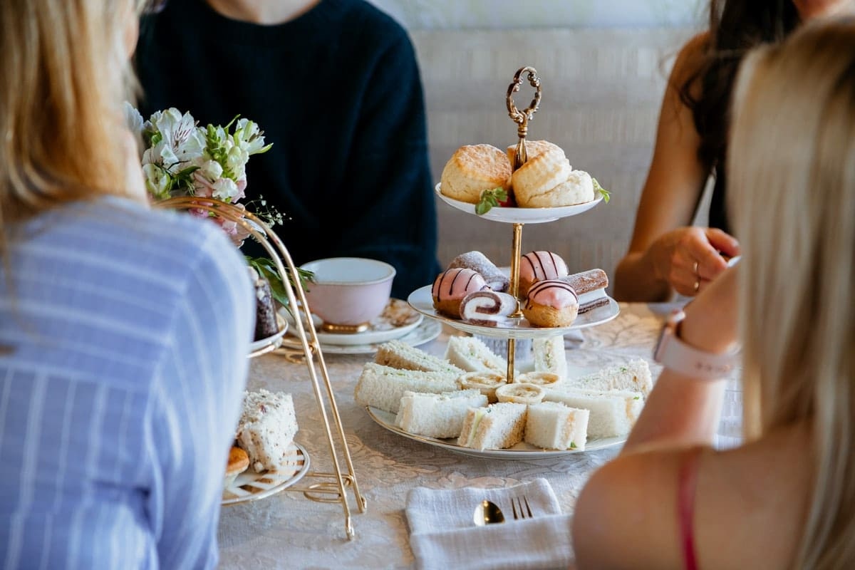 Close-up of three women gathered around gold three-tier afternoon tea stand displaying crustless finger sandwiches, pink and chocolate pastries with decorative toppings, and scones with strawberries on top tier, with white flower bouquet and teacups on rustic table