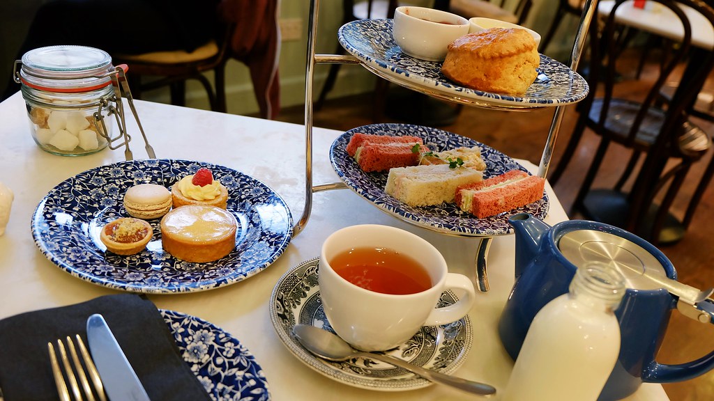 Afternoon tea service on white tablecloth featuring two-tier chrome stand with blue and white patterned plates, finger sandwiches and pink loaf cake slices on middle tier, scone on top tier, separate plate with pastries including lemon tart and macaron, white teacup with amber tea on silver saucer, navy blue teapot, glass jar with preserves