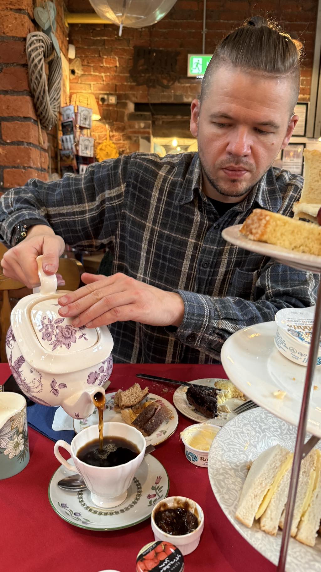 Person in black plaid flannel shirt pouring tea from purple floral teapot into white teacup on saucer at table with red tablecloth, afternoon tea spread with finger sandwiches, brownies, Victoria sponge cake on plates, jam pot, rustic exposed brick café interior