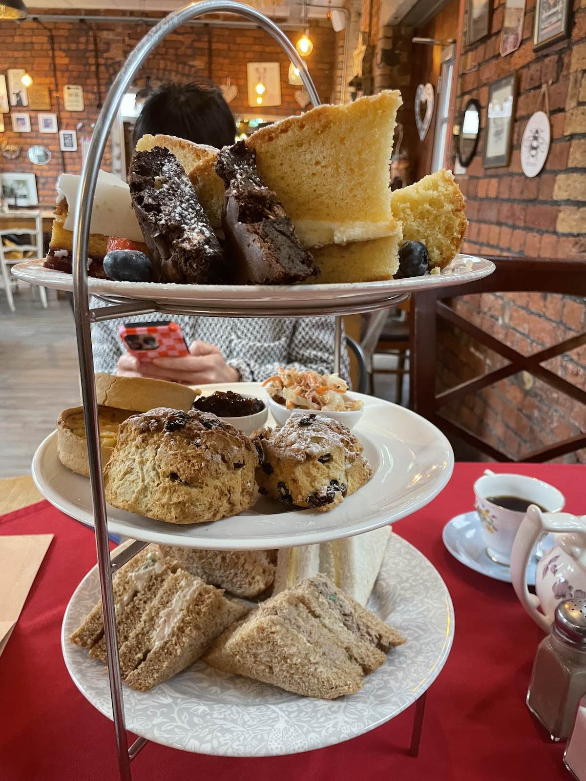 Three-tier chrome afternoon tea stand in rustic brick café with exposed beams, featuring finger sandwiches on bottom tier, scones with jam and coleslaw on middle tier, chocolate brownies with powdered sugar and Victoria sponge cake slices with blueberries on top tier, person holding phone, red tablecloth, teacups and teapot visible
