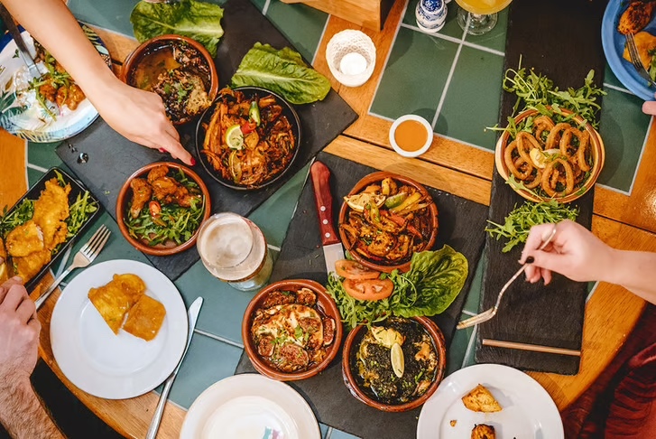 Overhead view of a shared dining table with multiple Korean dishes in brown stone bowls, including bibimbap and stir-fried vegetables, alongside lettuce wraps, crispy spring rolls, onion rings, and beer glasses on a green tiled table